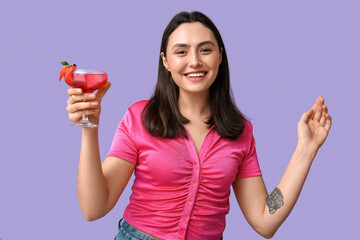 Beautiful young woman holding glass of tasty cocktail with strawberry on lilac background