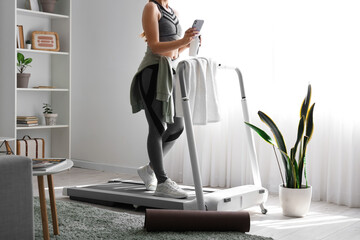 Sporty woman with mobile phone and water bottle training on treadmill at home