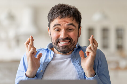 A man with short, dark hair and a beard sits indoors and crosses his fingers while looking at a laptop. He is wearing a blue shirt and has a hopeful expression on his face.