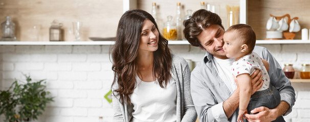 A young family smiles in their modern kitchen. The father holds their baby while the mother looks...