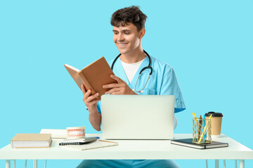 Male medical student reading book with modern laptop and jaw model at table on blue background