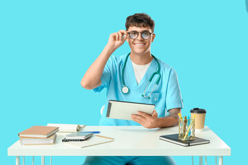 Male medical student with modern tablet computer, notebooks and paper cup of coffee at table on blue background