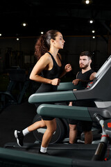 A male trainer motivates a brunette sportswoman as they run together on a treadmill in a gym setting.