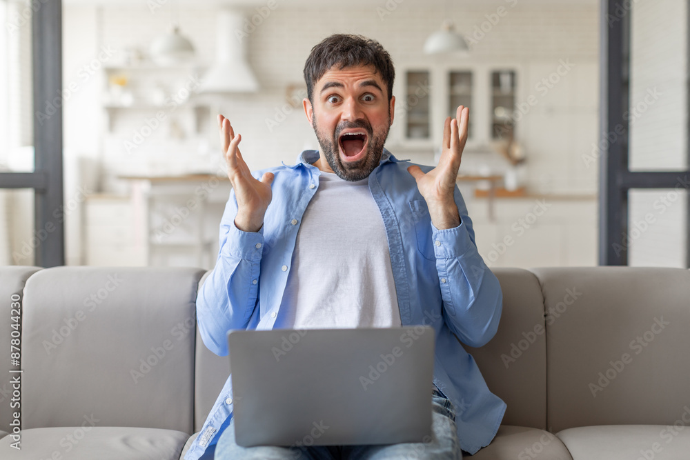 Poster A man with a shocked expression sits on a couch in his home with a laptop in his lap. He is surprised and excited by something on the screen.