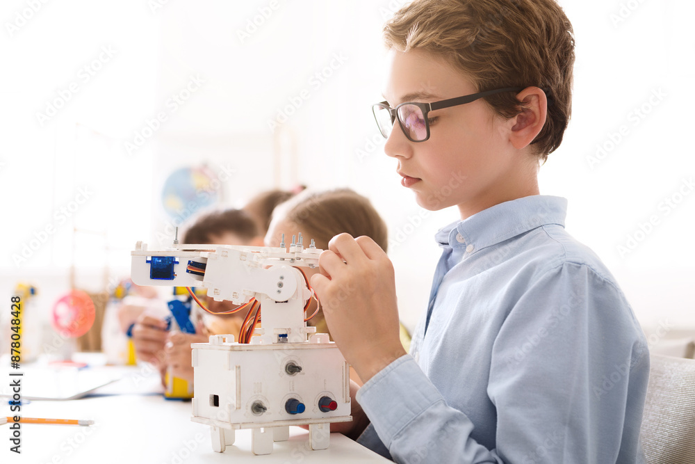 Poster A young boy with glasses is sitting at a table in a classroom setting and is working on building a small, white robotic arm. He is holding a small piece of the robot in his hand.