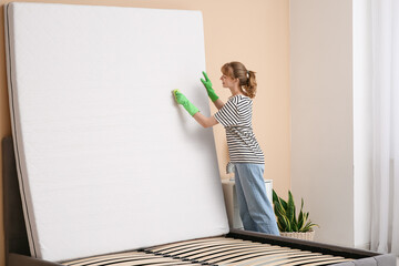 Young woman cleaning mattress with brush in bedroom
