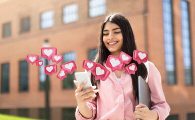 A young woman with long, dark hair is smiling while looking at her smartphone. She is standing in...