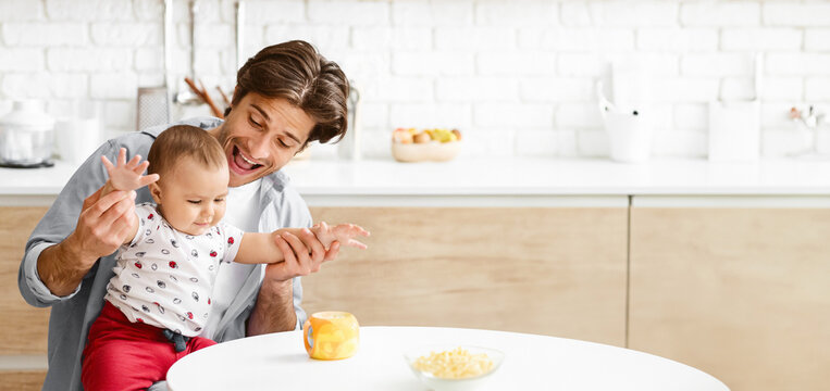 This image shows a father and his young son playing in a kitchen. The father is holding his son, who is reaching out with his hand and smiling. The father is also smiling, copy space