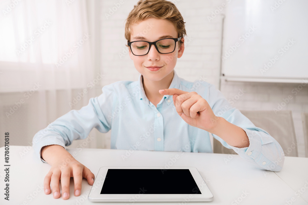 Poster This image shows a young boy wearing glasses sitting at a white table. He is using a tablet and is pointing at the screen with his index finger. He appears to be focused on the device