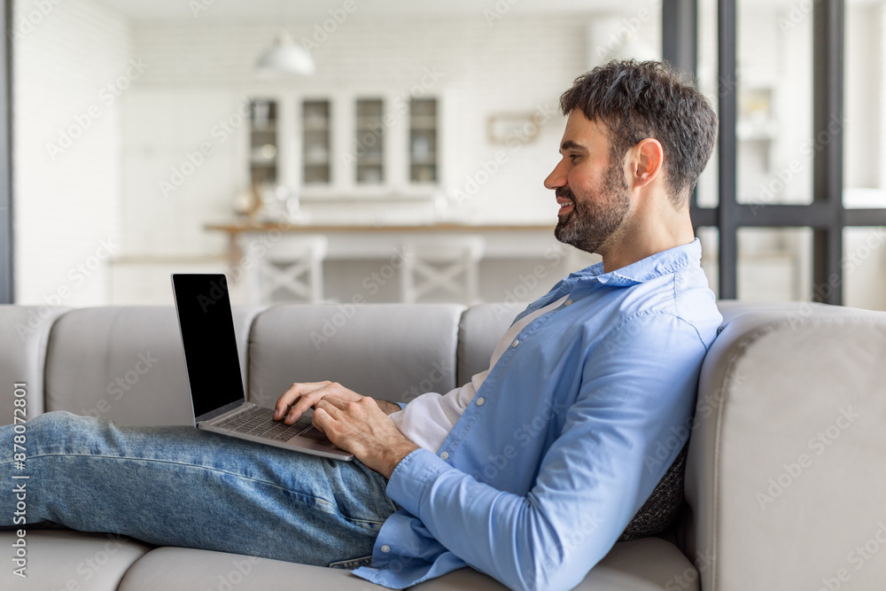 Poster this image shows a man relaxing on a gray sofa, using a laptop. he is wearing jeans and a light blue