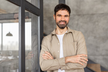 Obraz premium A man stands with his arms crossed, leaning against a glass door in a modern business setting. He is wearing a light brown button-up shirt over a white t-shirt.