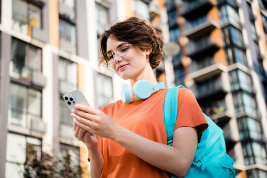 Photo of positive charming lady wear orange t-shirt spectacles rucksack chatting apple samsung iphone modern device outside urban city street