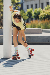 Young woman in yellow crop top and brown shorts balances herself on roller skates, holding onto pole for support. Concept of sport, active lifestyle, youth, summer holidays, hobby.