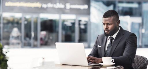 Fototapeta premium Serious African American Businessman Working On Laptop Computer In Outdoor Cafe In City Center. Free Space