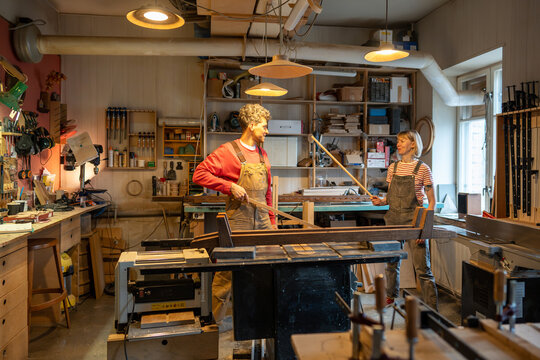 Joyful man, woman jokingly fighting, using wooden sticks as swords. Happy unserious girl, guy in carpentry studio idle, slack off from work process, taking break. Lazy careless spouses joiners. 