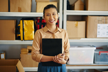 Portrait of Asian female worker with document smiling at camera while standing in warehouse