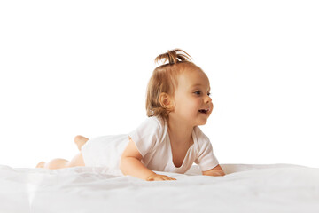 Fototapeta premium Happy little baby girl, child in bodysuit ling on belly in bed and making moves, crawling isolated on white background. Concept of childhood, care, health, infancy, lifestyle, motherhood