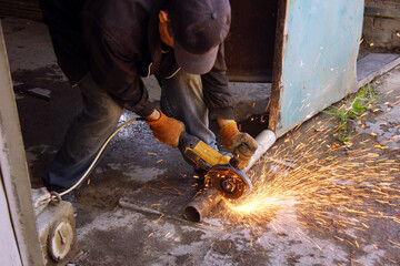 A man wearing gloves and a cap uses a grinding tool to cut a metal pipe outside. Sparks fly as he works. The pipe is resting on the ground in front of an open door.