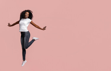 Fototapeta premium A young Black woman with curly hair is jumping in front of a pink background. She is wearing a white t-shirt and blue jeans. She is smiling and her arms are outstretched.