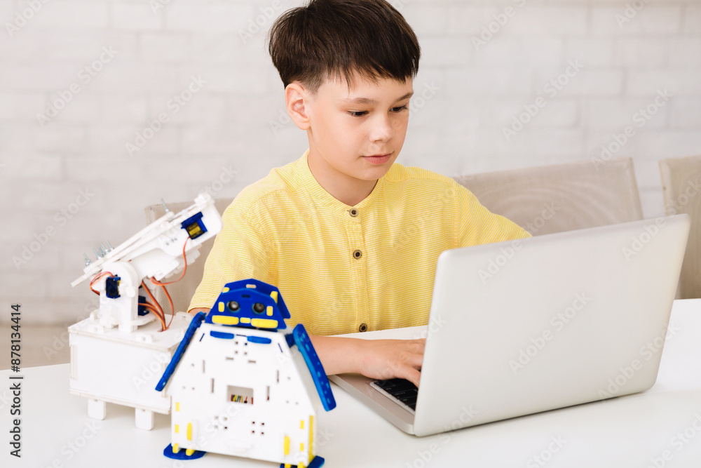 Poster a young boy is sitting at a desk, using a laptop to program a small robot. the robot is made of whit