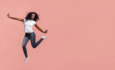 This image shows a young African American woman with dark hair wearing a white shirt and blue jeans jumping in front of a pink background. She is smiling and appears to be enjoying herself.