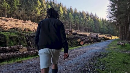 black male hiking on trail up peaceful lush high elevation green forest with sun flares tracking shot from behind. Black Man hiking and exploring forest area in Dublin Mountains - Ireland