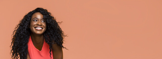 The image shows a young black woman with curly black hair smiling and looking upwards. She is wearing a red tank top and has a natural, healthy look. The background is a solid peach color.