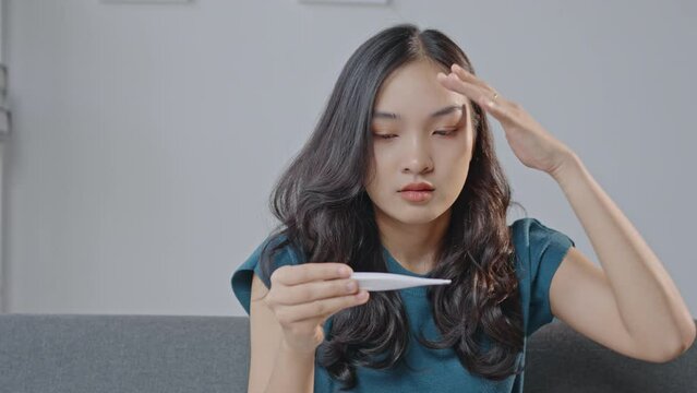 Young woman sitting on couch, checking thermometer and feeling unwell, touching forehead and neck with worried expression