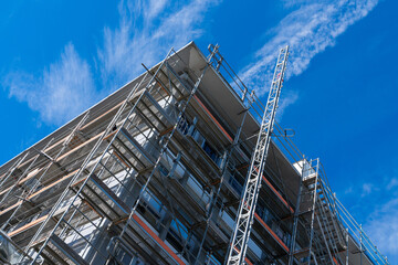 A tall apartment building under construction, with scaffolding covering the exterior. A clear blue sky with wispy clouds.