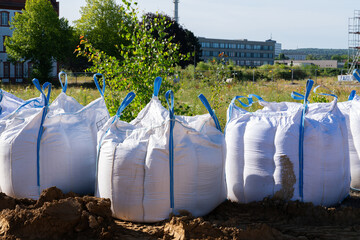 Several large white bags sit on a dirt construction site. Buildings and trees can be seen in the...