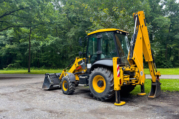 A yellow backhoe is parked on a gravel path in a forest setting. Its backhoe arm is raised and its bucket is resting on the ground.