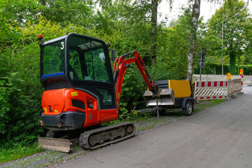 A red mini excavator is parked next to a trailer, both near a forested area.