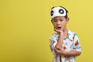 Portrait of Asian child sleepy and yawning, wearing pajamas and sleep mask, putting his hand under his cheek isolated on yellow background. Healthy sleep concept. Copy space
