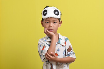 Portrait of sleepy Asian child wearing pajamas and sleep mask isolated on yellow background. Child wants to sleep. Healthy sleep concept. Copy space