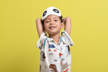 Portrait of Asian child stretching arms, wearing pajamas and sleep mask isolated on yellow background. Child wants to sleep. Healthy sleep concept. Copy space