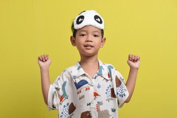 Portrait of cheerful Asian child doing stretching. Child wearing pajamas and sleep mask isolated on yellow background. Child wants to sleep. Healthy sleep concept. Copy space