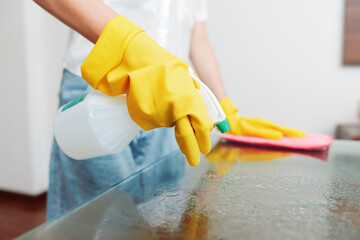 Woman in yellow rubber gloves cleaning glass table with spray bottle and yellow glove on the other side