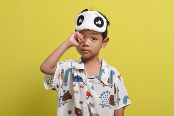 Portrait of sleepy Asian child rubbing his eyes isolated on yellow background. Child wearing pajamas and sleep mask. Healthy sleep concept. Copy space