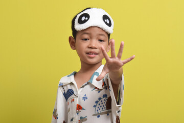 Portrait of Asian child wearing pajamas and sleeping mask isolated on yellow background. Child wants to sleep. Healthy sleep concept. Copy space