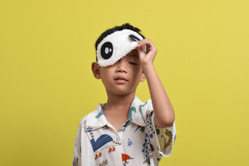 Portrait of cute Asian child taking off sleeping mask isolated on yellow background. wearing pajamas. Healthy sleep concept. Copy space