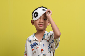 Portrait of cute Asian child taking off sleeping mask isolated on yellow background. wearing pajamas. Healthy sleep concept. Copy space