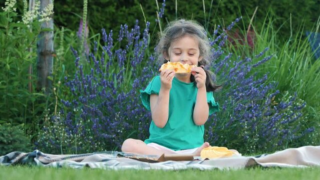 A happy girl eagerly eating a slice of pizza while sitting on a blanket at an impromptu picnic.