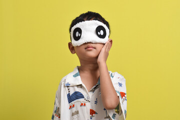 Portrait of Asian child wearing pajamas and sleeping mask isolated on yellow background. Child wants to sleep. Healthy sleep concept. Copy space