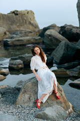 Serene woman in white dress sitting on ocean rock with vibrant red shoes, contemplating the horizon