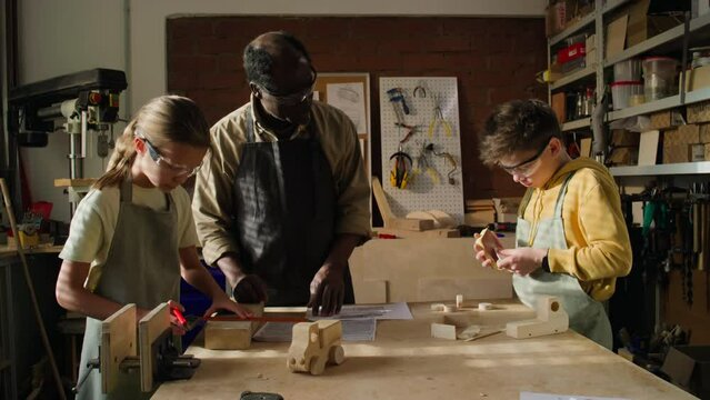 Little Caucasian kids wearing protective glasses sanding wooden details with assistance of mature Black teacher during woodworking class