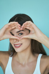 Young Woman Making Heart Shape With Hands Around Eyes, Smiling, And Looking At Camera Against Blue Background.