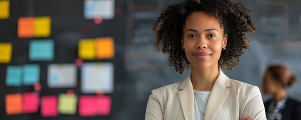 A woman with curly hair is standing in front of a board with colorful sticky notes. She is wearing a white jacket and smiling