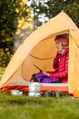 A woman sits in a tent with a tablet in front of her. She is wearing a red jacket and a pink hat. The tent is orange and has a small opening. The scene is peaceful and serene