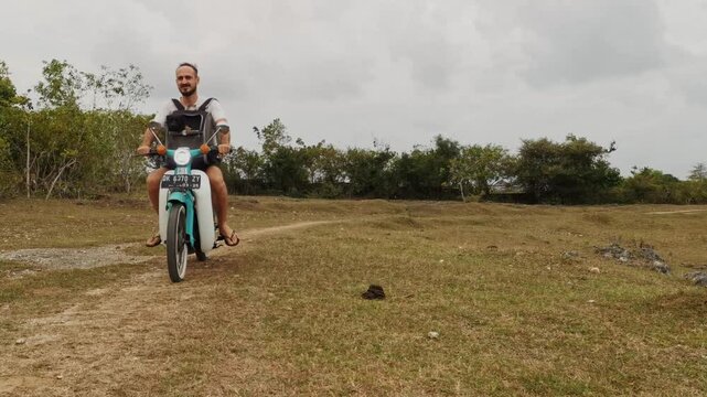 Steady shot of a European man cruising on a white and turquoise Honda C70 motorcycle along a gravel path on a cloudy day. Man carries a front bag with a small black dog. Practical adventure footage.