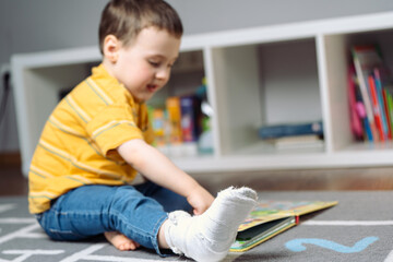 Toddler boy with a bandage or cast on his leg plays with colourful book. Fracture of a foot and finger in children. Human healthcare and medicine concept. focus on the cast